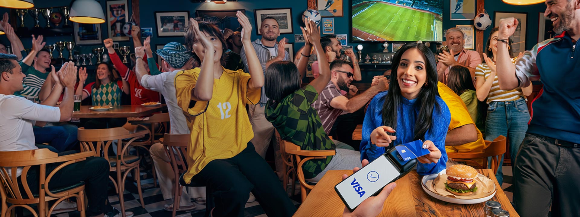A woman holds a phone with a Visa payment app in a lively sports bar, surrounded by cheering fans watching a soccer match on TV.