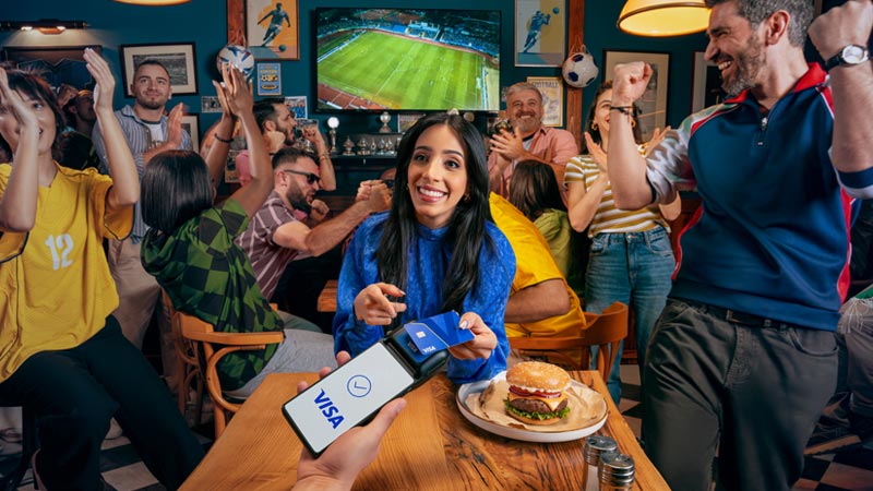 A woman holds a phone with a Visa payment app in a lively sports bar, surrounded by cheering fans watching a soccer match on TV.