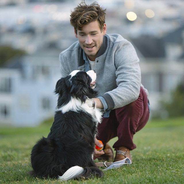 Man kneeling on a hillside grass area petting a dog.