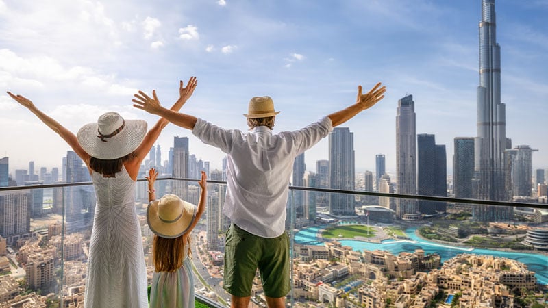 Three people in hats stand on a balcony with arms raised, overlooking a modern city skyline with tall skyscrapers on a sunny day.