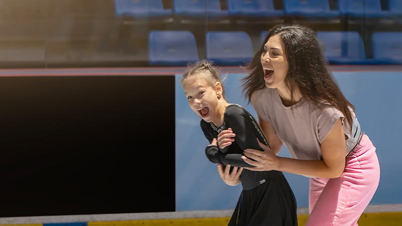 A woman helps a young girl in a black outfit ice skate; both are smiling widely inside an indoor ice rink.