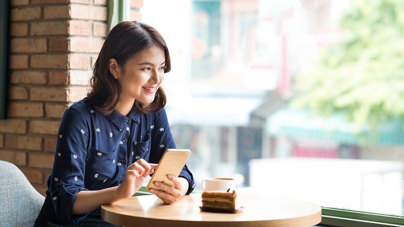 Woman sitting at a café table by a window, holding a smartphone, with a cup of coffee and a slice of cake in front of her.