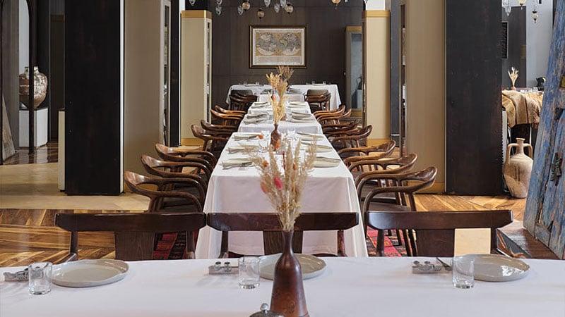 A long dining table covered with a white tablecloth, set with plates, glasses, and cutlery, surrounded by wooden chairs and decorated with a vase of dried flowers.
