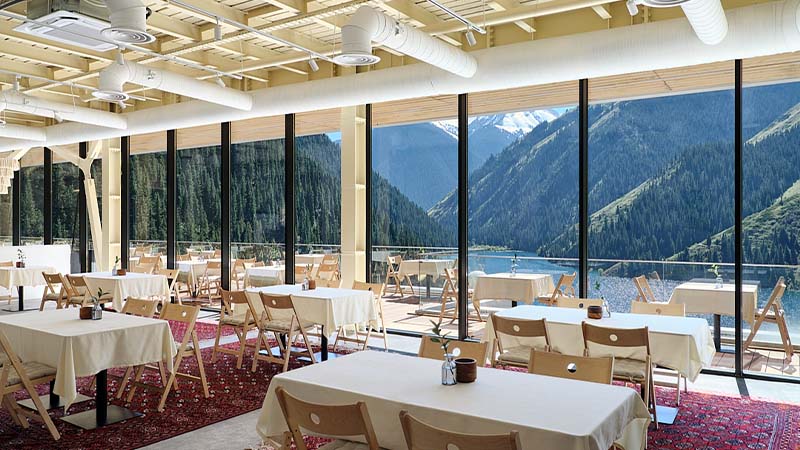 A dining area with tables and chairs features floor-to-ceiling windows overlooking mountains, forests, and a lake on a sunny day.