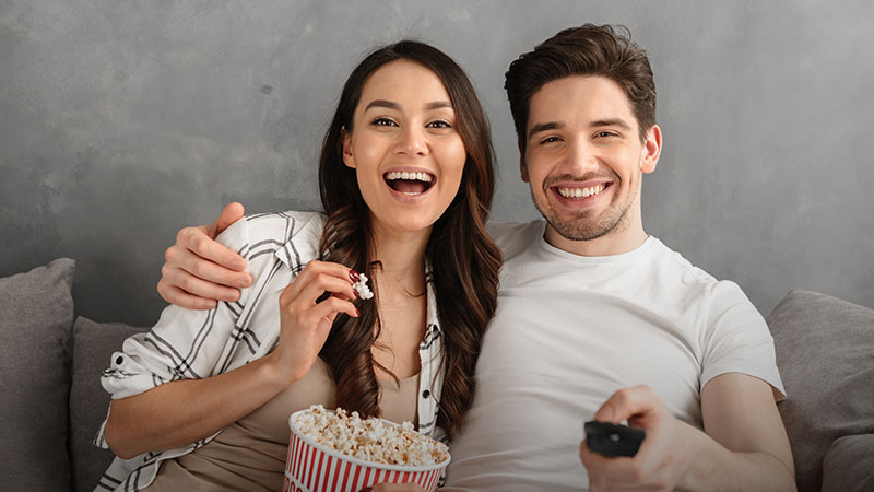 A man and a woman watching TV and eating popcorn